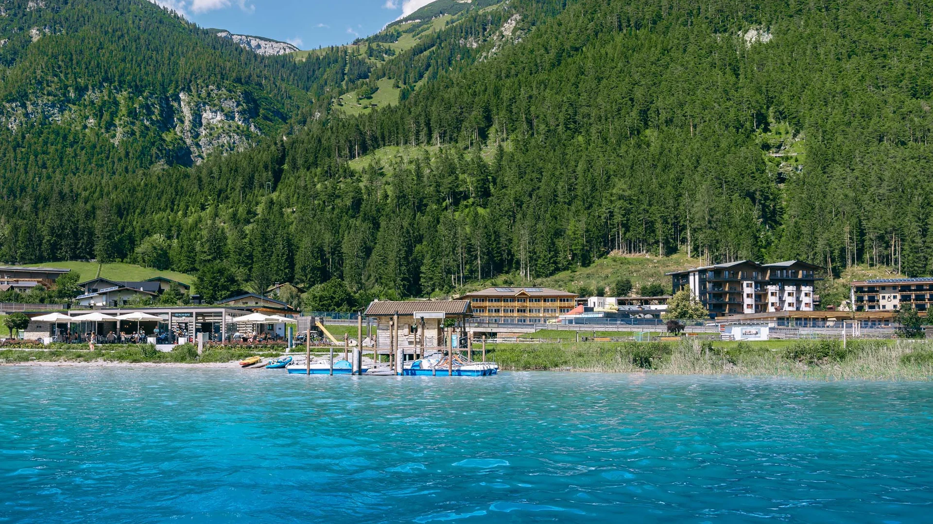 Klares blaues Wasser mit Bergkulisse und Gebäude am Ufer bei sonnigem Wetter