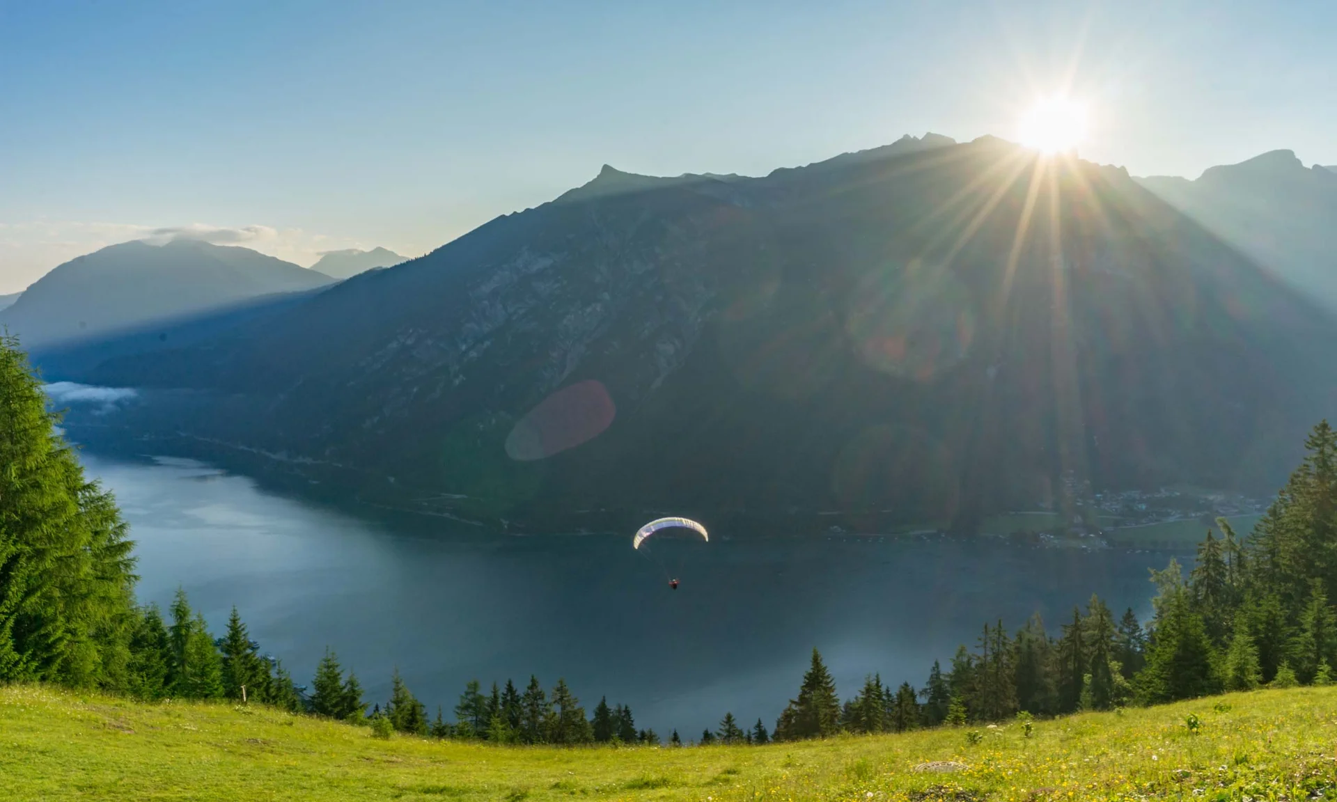 Paraglider fliegt über See mit Bergen und Sonnenuntergang im Hintergrund