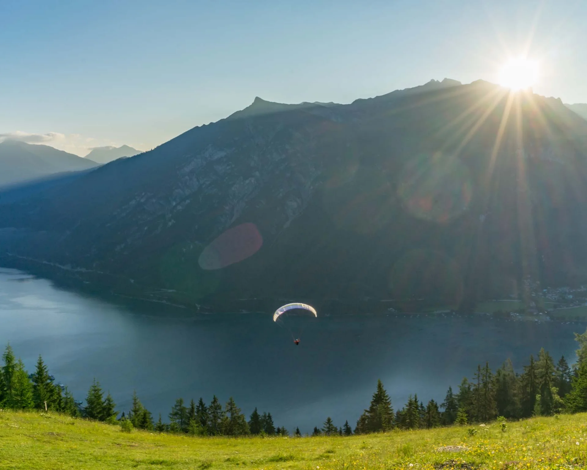 Paraglider fliegt über See mit Bergen und Sonnenuntergang im Hintergrund