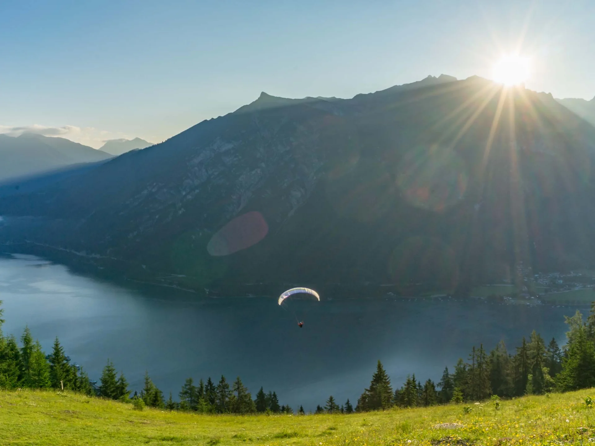 Paraglider fliegt über See mit Bergen und Sonnenuntergang im Hintergrund
