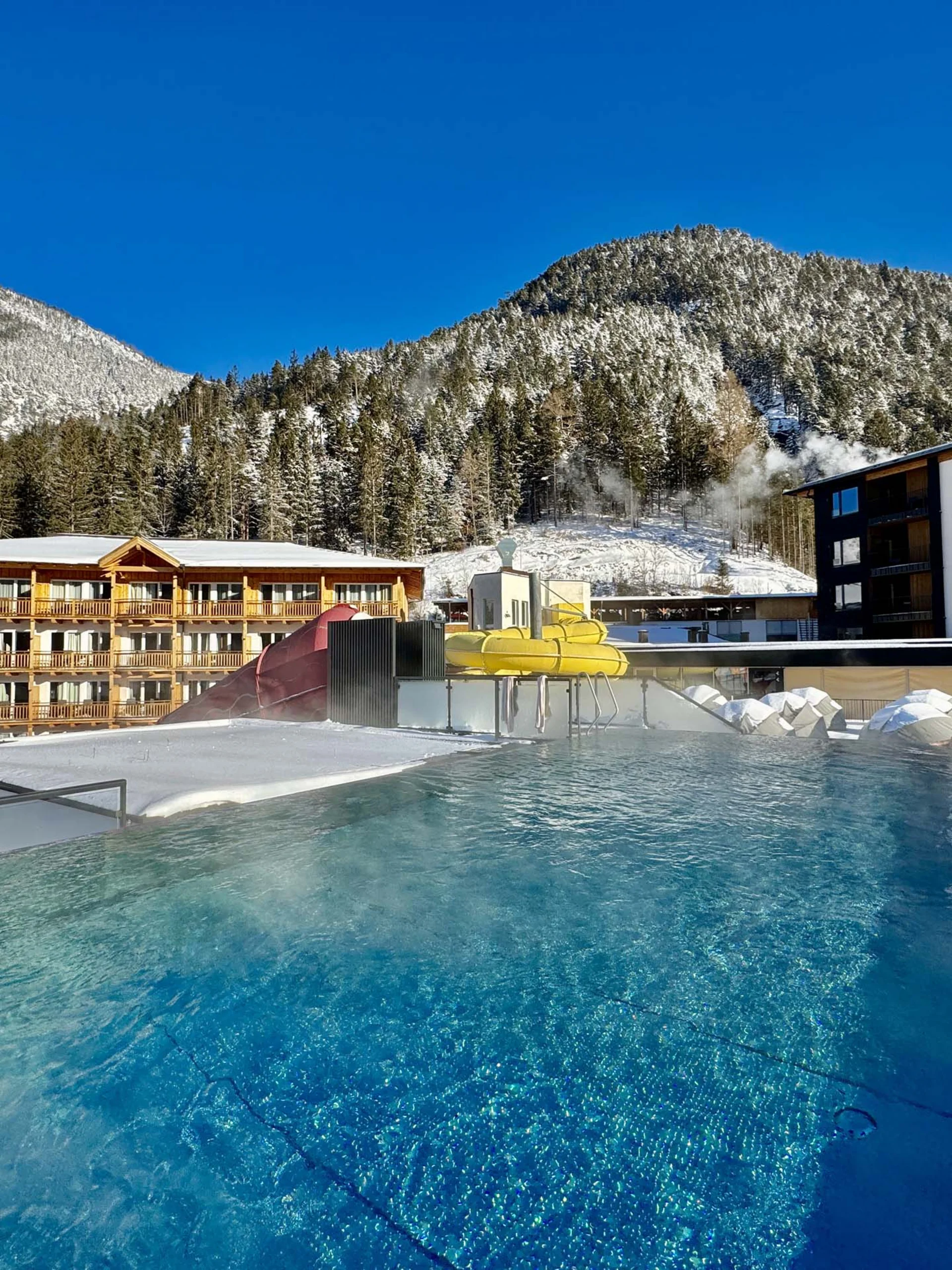 Beheizter Außenpool mit Wasserrutsche in verschneiter Berglandschaft bei klarem Himmel