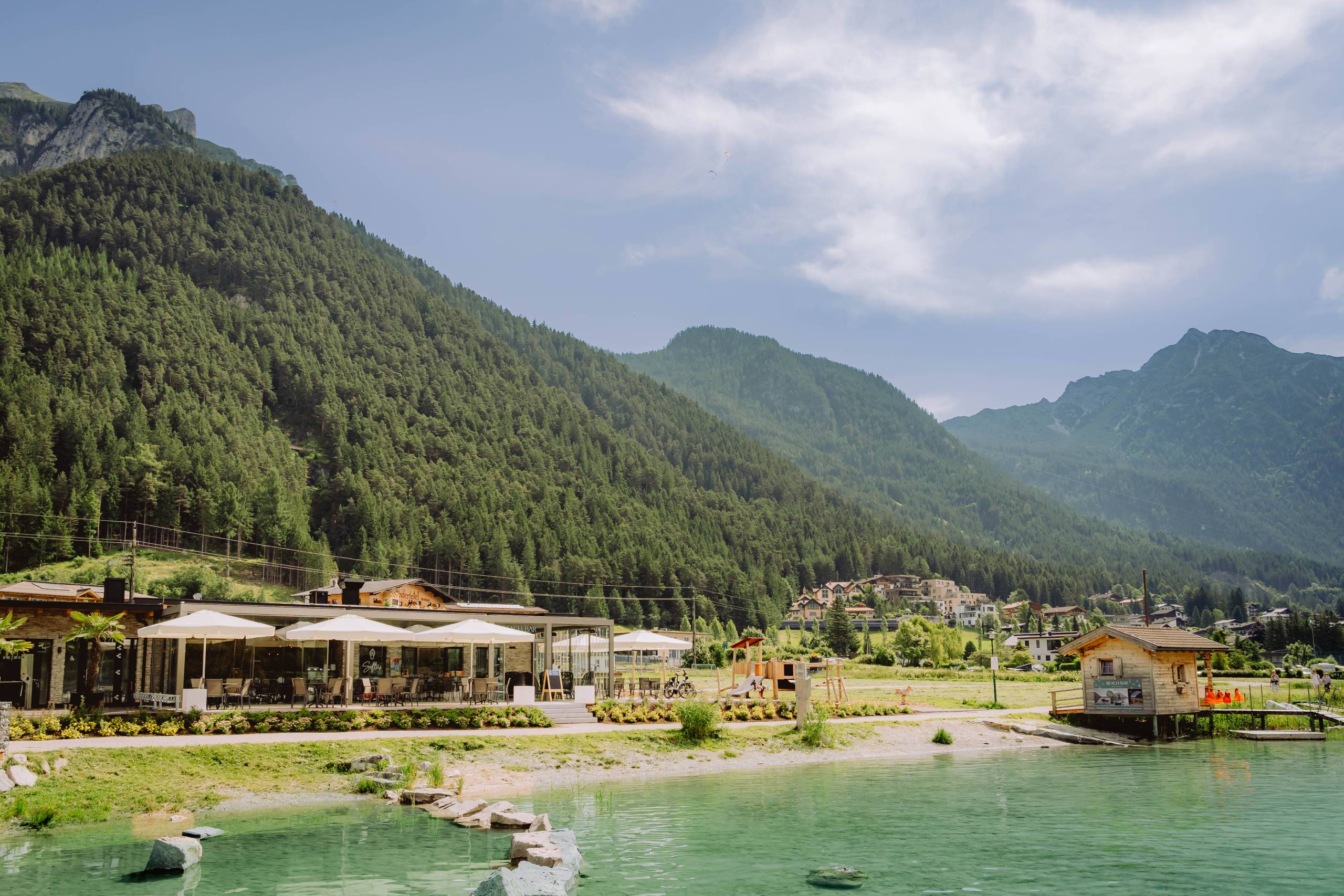 See mit Restaurant, Spielplatz und Berghütten vor bewaldeten Bergen