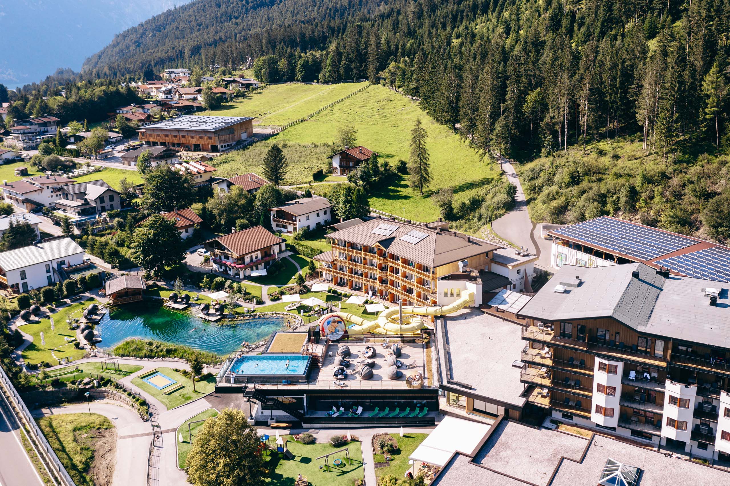 Luftaufnahme eines Resorts mit Swimmingpools und Wasserrutsche in grüner Alpenlandschaft